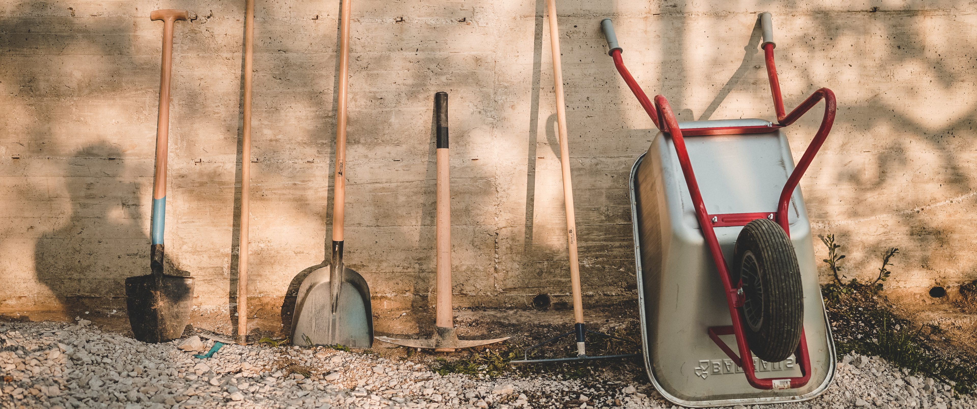 wooden spades, pickaxe, rake and silver red wheelbarrow leaned against a sun-dappled concrete wall